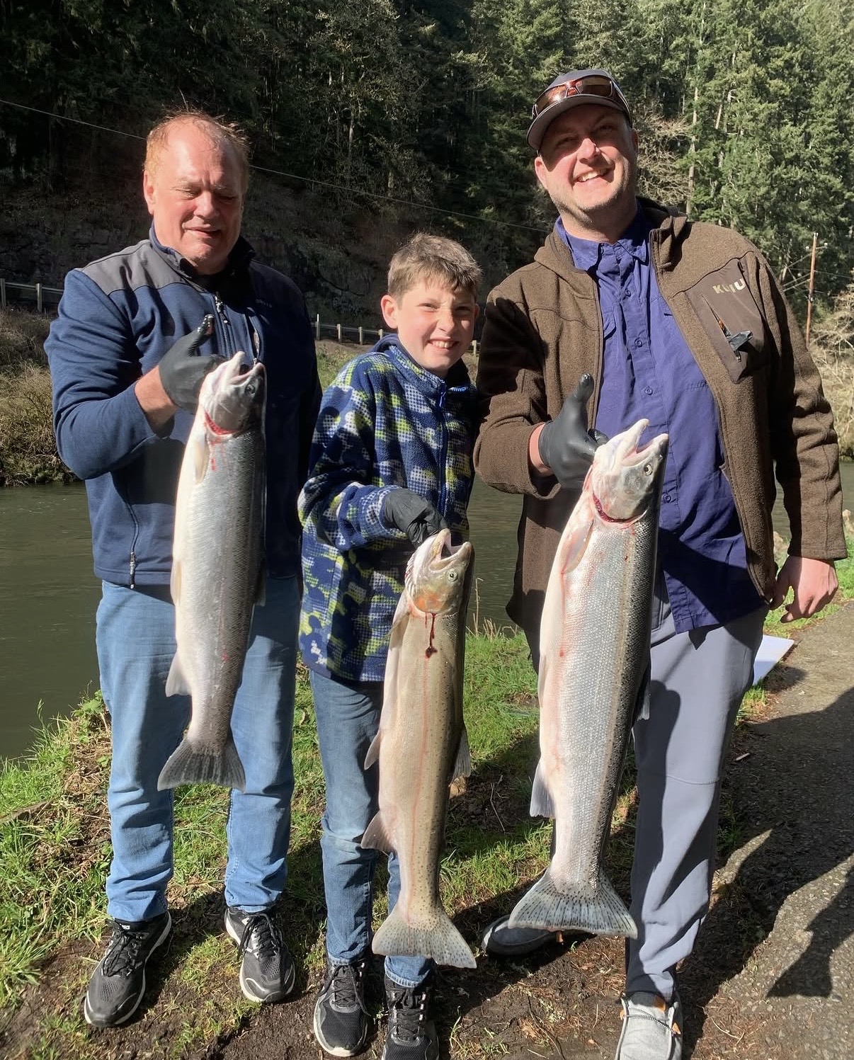 Three anglers, including a young boy, each holding a bright steelhead beside the river