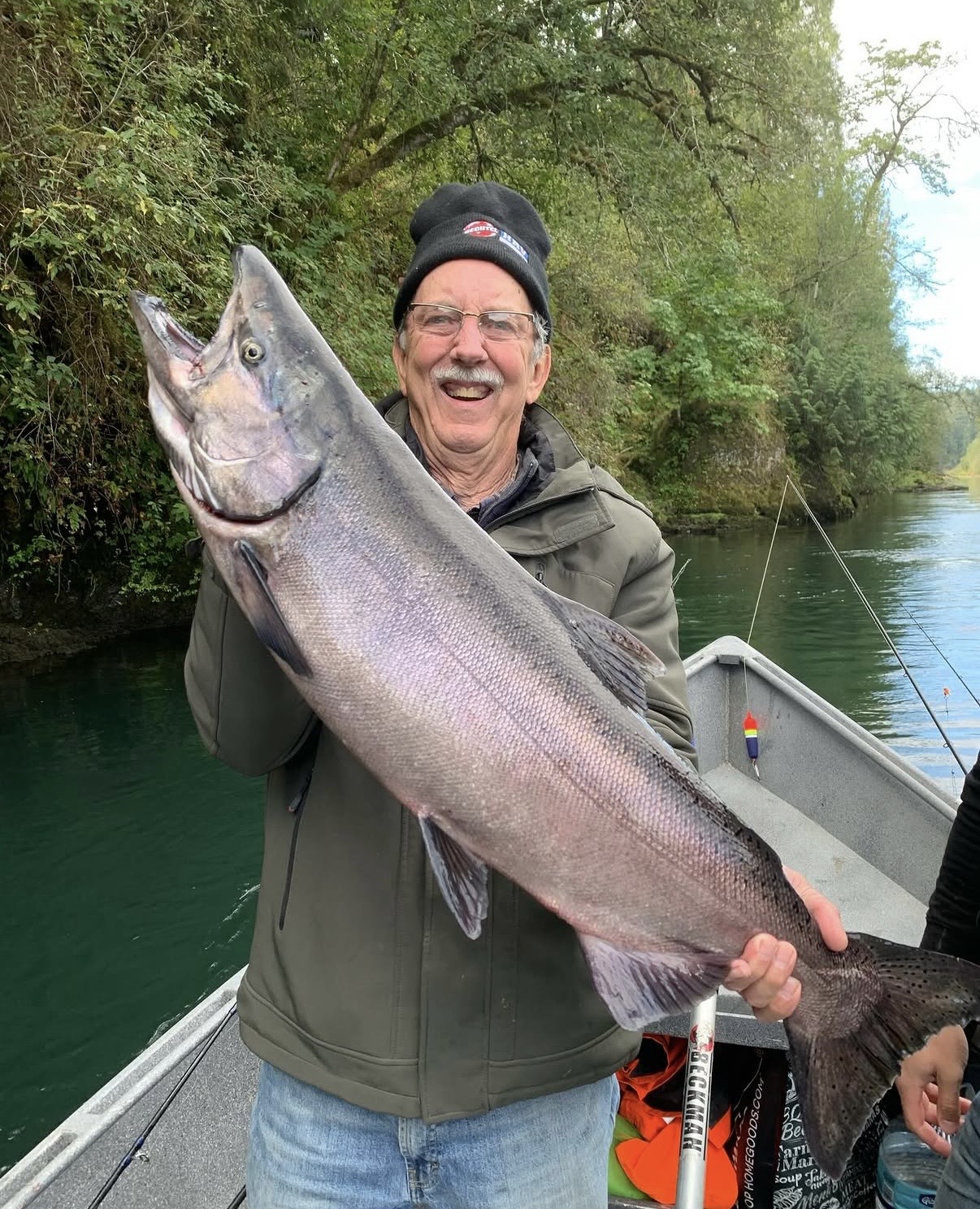 Happy angler in a beanie holding a big chrome Chinook salmon on a green, tree-lined river